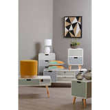 Milo Chest of 4 Drawers featuring a modern design with a light wood finish, four spacious drawers with sleek handles, and tapered legs. The chest is positioned in a stylish living space alongside a yellow accent pillow, a small lamp, and decorative items on top. A geometric wall art piece is visible in the background, enhancing the contemporary aesthetic