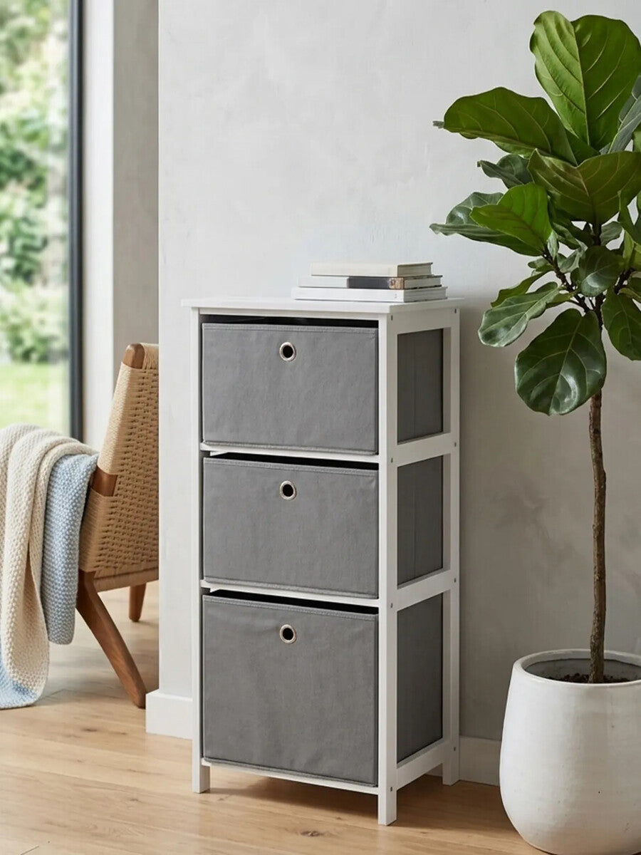 Three grey fabric drawers in a cabinet with a white frame, positioned next to a large window with natural light. A green potted plant is nearby, and a cozy chair with a throw blanket is partially visible in the background