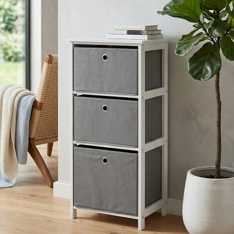 Three grey fabric drawers in a white cabinet with a modern design, placed against a light-colored wall. A potted plant is visible beside the cabinet, and a cozy throw blanket is draped over a nearby chair. The floor is wooden, adding warmth to the scene