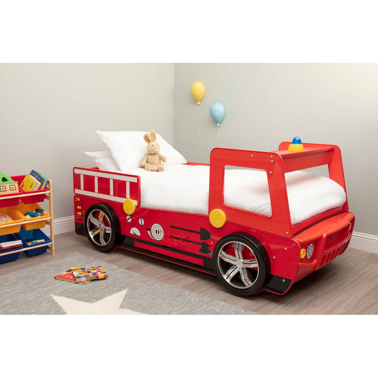 A vibrant red fire engine bed designed for kids, featuring a detailed front with wheels and a ladder. The bed is positioned in a playful room with a soft white mattress, a teddy bear on top, and colorful balloons in the background. A toy storage unit filled with various toys is visible to the left, along with scattered building blocks on the floor