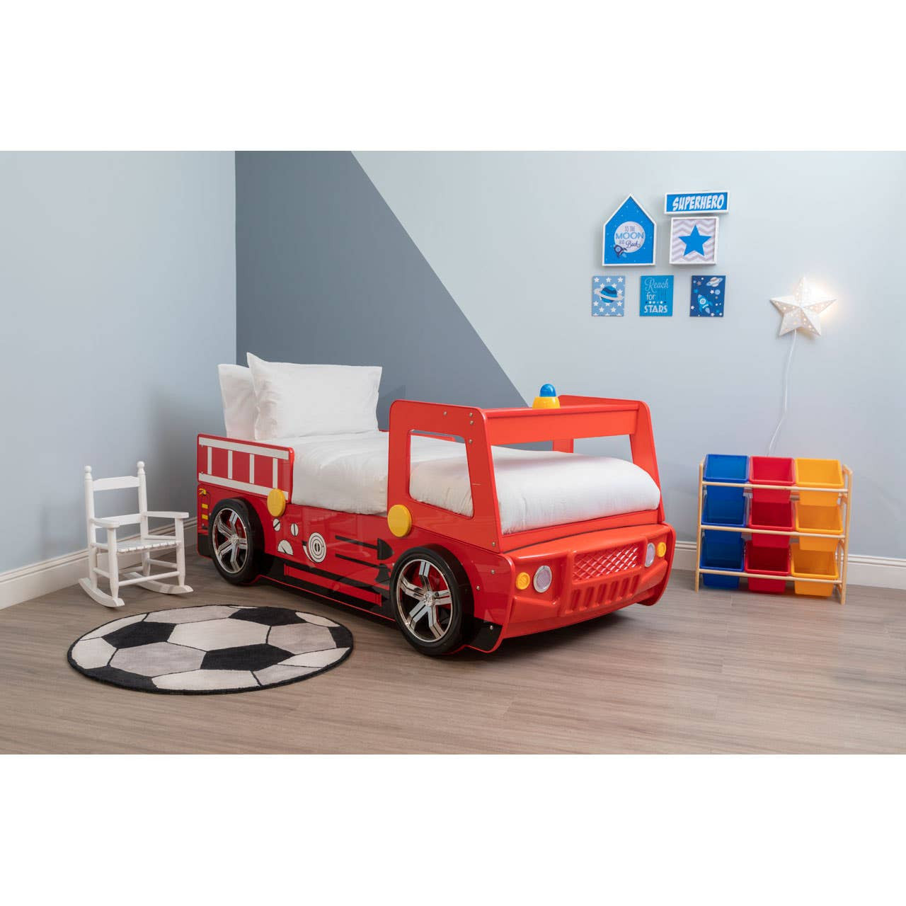 A vibrant red fire engine bed designed for kids, featuring large wheels and a playful design. The bed is positioned in a bright room with light blue walls, a circular black and white rug on the floor, a small white chair, and colorful storage bins stacked nearby. Wall decals in the shape of a house and various shapes add a fun touch to the decor