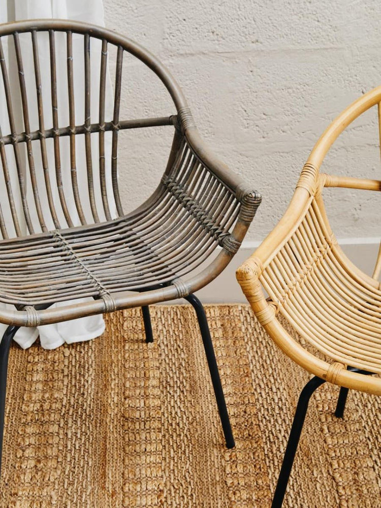 A grey wash natural rattan chair with a curved backrest and open design, positioned next to a lighter rattan chair, both resting on a textured woven rug. The background features a neutral wall, enhancing the natural tones of the chairs