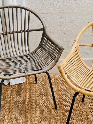A grey wash natural rattan chair with a curved backrest and open design, positioned next to a lighter rattan chair, both resting on a textured woven rug. The background features a neutral wall, enhancing the natural tones of the chairs