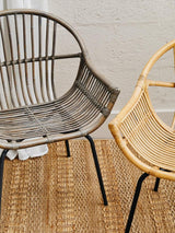 A grey wash natural rattan chair with a curved backrest and open design, positioned next to a lighter rattan chair, both resting on a textured woven rug. The background features a neutral wall, enhancing the natural tones of the chairs