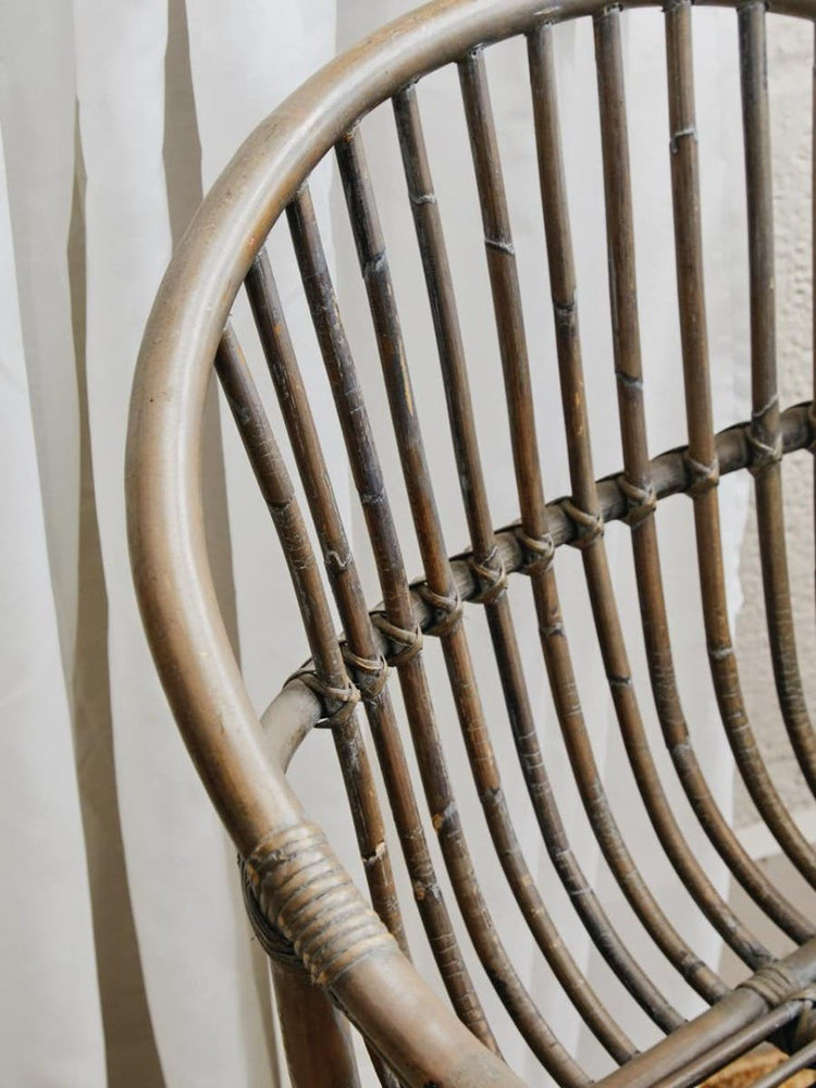 A close-up view of a grey wash natural rattan chair featuring a curved backrest with vertical slats, showcasing the intricate weaving of the rattan material against a soft, neutral background