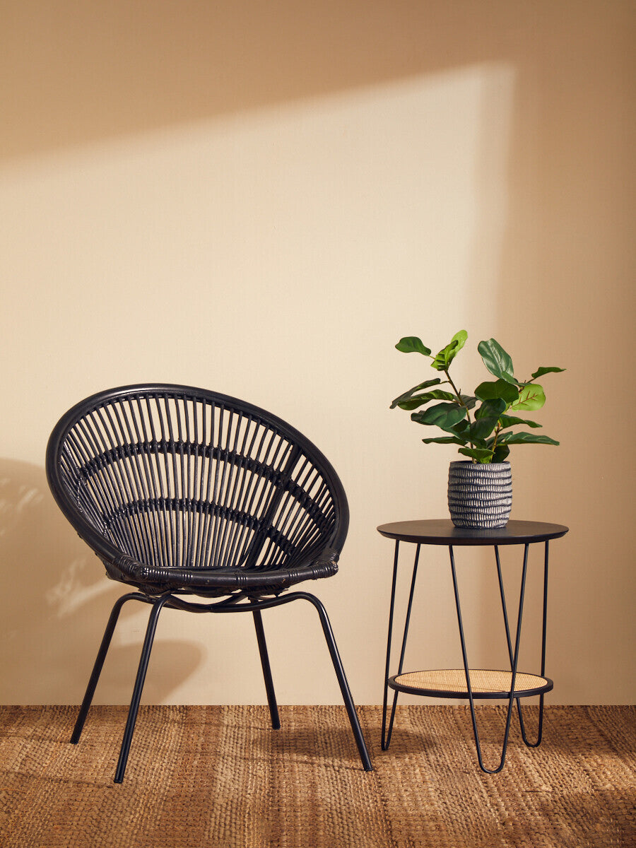 A black natural rattan chair with a curved backrest and slender iron legs is positioned next to a small round table. The table holds a potted plant, and soft sunlight casts a warm glow on the wall behind, creating a cozy atmosphere. The floor is made of wooden planks, enhancing the natural aesthetic of the scene