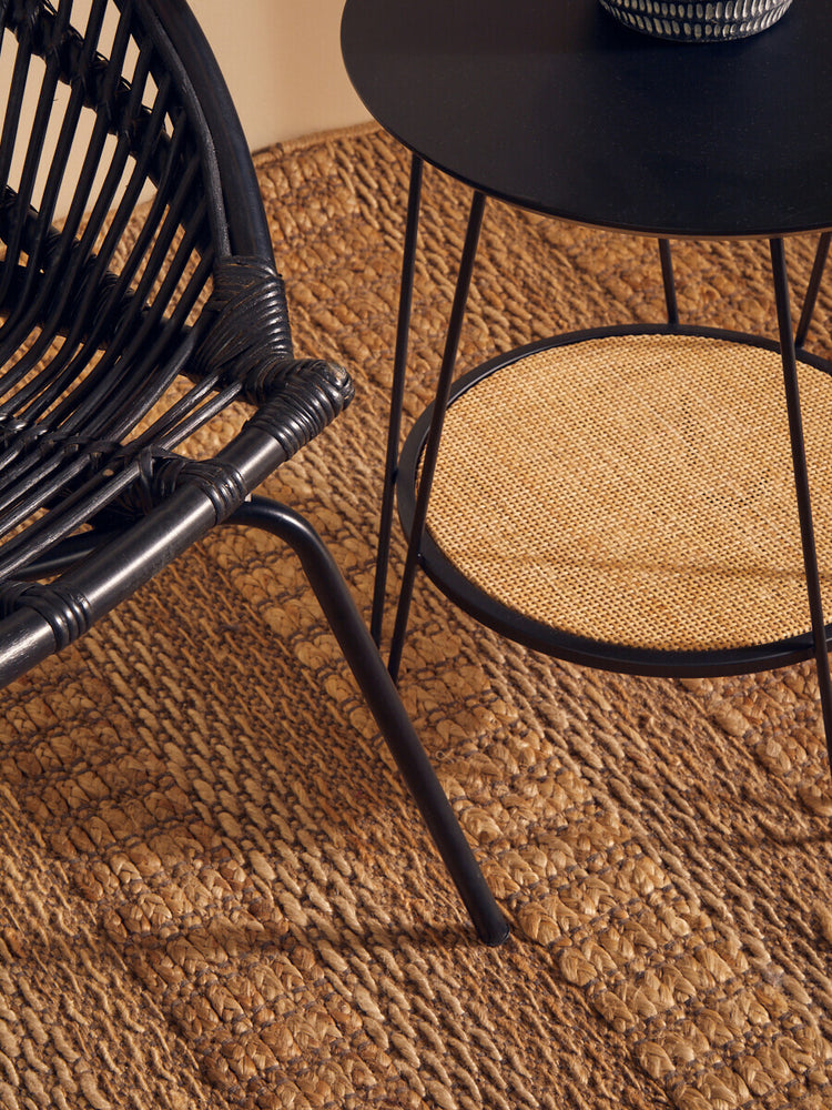 A black natural rattan chair with a curved backrest and iron legs is positioned next to a small black side table. The chair features a woven design, and the floor is covered with a textured natural fiber rug. A potted plant sits on the table, adding a touch of greenery to the scene