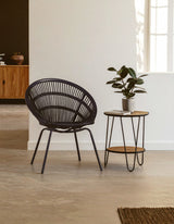 A black natural rattan chair with a curved backrest and slender iron legs is positioned next to a small wooden side table. The table features a round top and thin metal legs, and a potted plant sits on it. The background includes a light-colored wall and a glimpse of wooden cabinetry. The flooring is a neutral tone, enhancing the modern aesthetic of the space