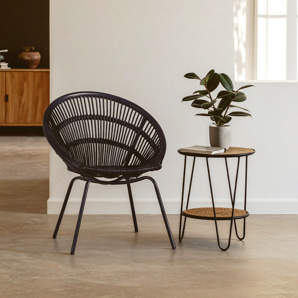 Black natural rattan chair with a curved backrest and iron legs, positioned next to a small round side table with a wooden top and metal base, featuring a potted plant on the table, set against a light-colored wall and a softly lit interior space