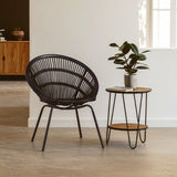 Black natural rattan chair with a curved backrest and iron legs, positioned next to a small round side table with a wooden top and metal base, featuring a potted plant on the table, set against a light-colored wall and a softly lit interior space