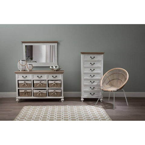 A white washed natural rattan chair positioned next to a tall white dresser and a low dresser with a mirror, all set against a soft green wall. A light-colored area rug is visible on the floor, adding warmth to the space