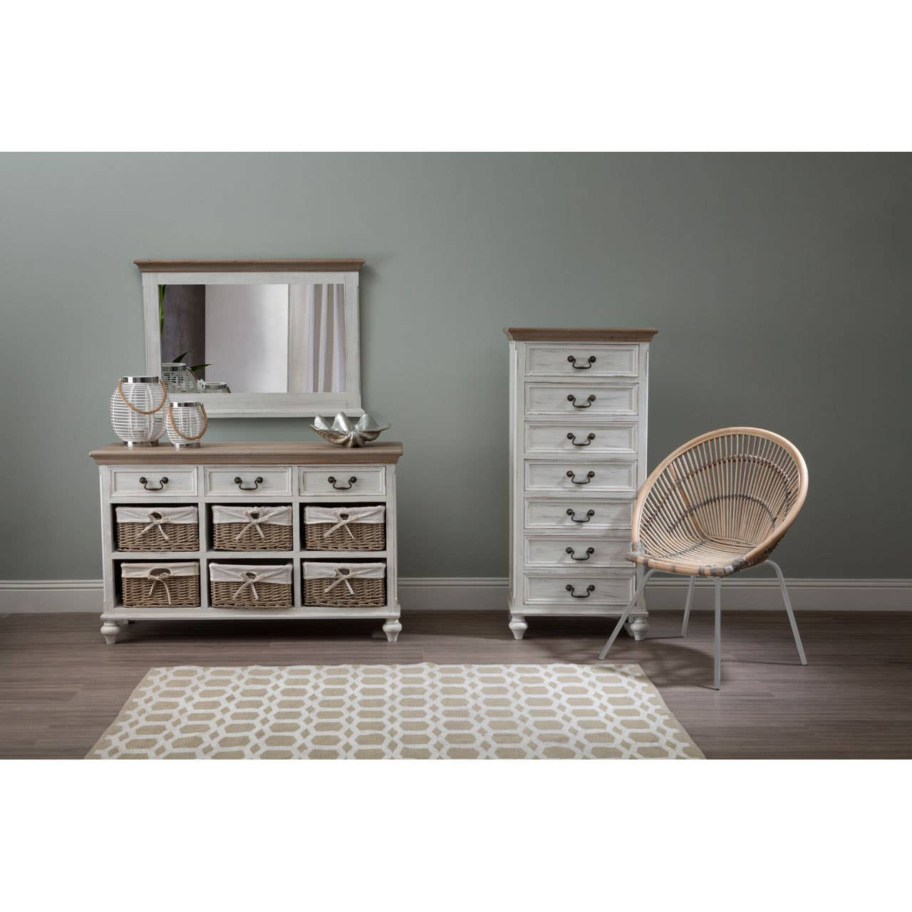 A white washed natural rattan chair positioned next to a tall white dresser and a low dresser with a mirror, all set against a soft green wall. A light-colored area rug is visible on the floor, adding warmth to the space