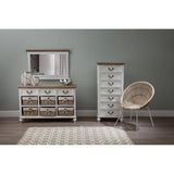 A white washed natural rattan chair positioned next to a tall white dresser and a low dresser with a mirror, all set against a soft green wall. A light-colored area rug is visible on the floor, adding warmth to the space