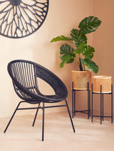 A black natural rattan and iron chair with a circular design, positioned next to two potted plants on a light-colored floor. The background features a decorative wall piece with a geometric pattern, and the overall setting has warm, neutral tones