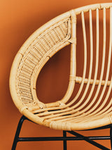 Natural rattan chair with a curved backrest and black iron legs, set against an orange background. The chair features a distinctive teardrop shape in the back, showcasing the woven rattan texture