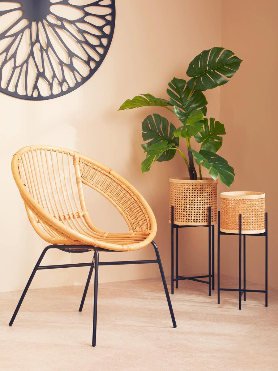 Natural rattan chair with a circular design and black iron legs, positioned next to a small plant and two wooden storage boxes on a black metal stand, set against a warm, neutral wall with a decorative wall hanging