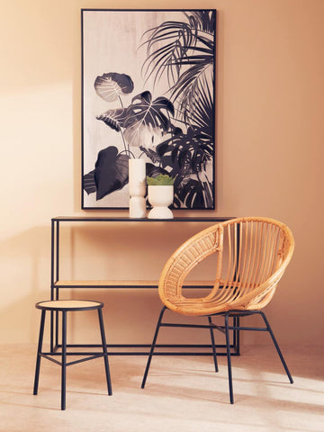 A natural rattan chair with a curved backrest and black iron legs is positioned next to a small black iron table. The scene features a large framed artwork with tropical leaves on a peach-colored wall, accompanied by two candles and a small plant on the table. A second stool is visible beside the chair