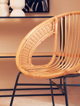 Natural rattan chair with a curved backrest and black iron legs, showcasing a modern design. The chair is positioned near a shelf with decorative items, set against a warm-toned wall