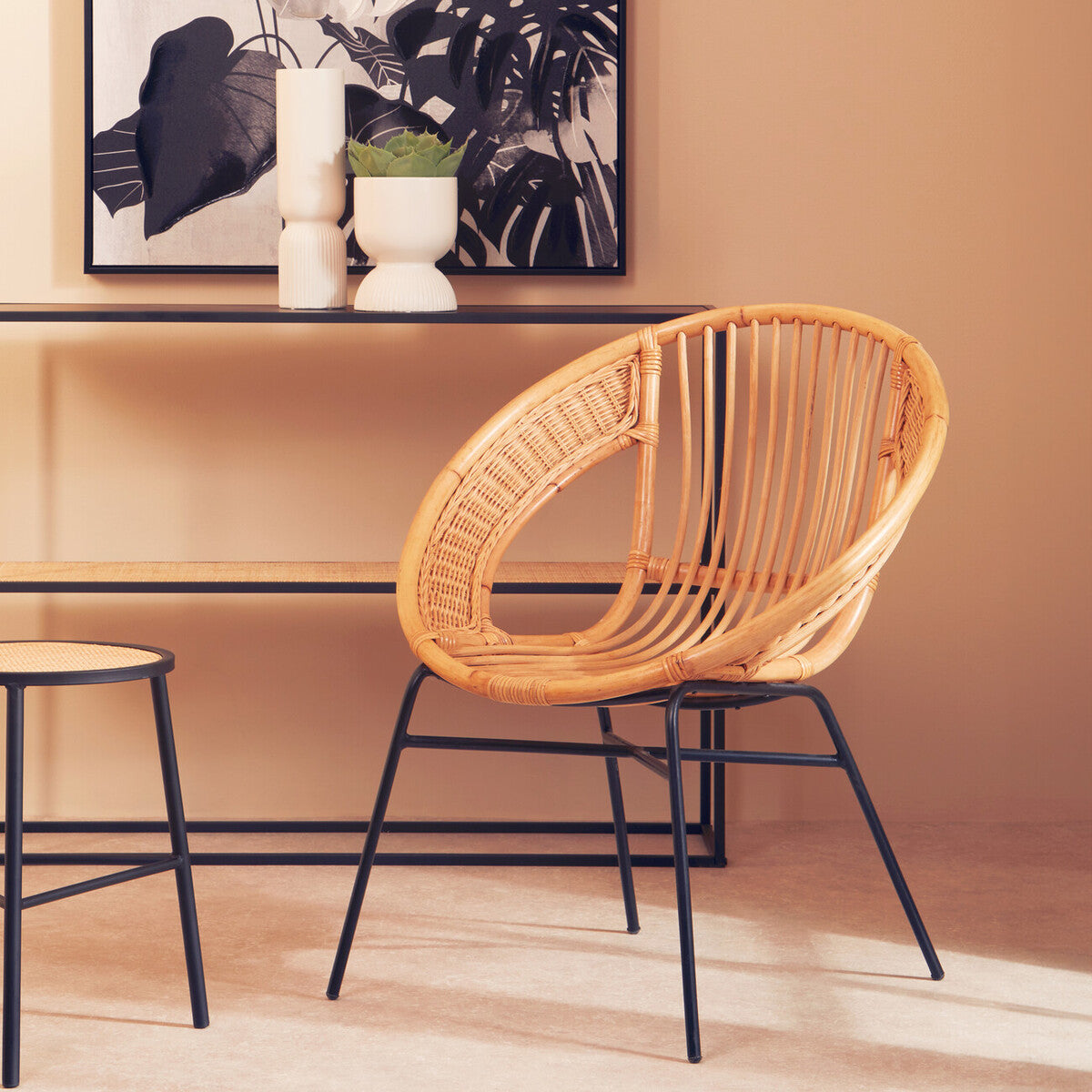 A natural rattan chair with a circular design and black iron legs, positioned next to a minimalist table. The chair features a woven texture and is set against a warm, neutral wall, with a decorative piece and a framed artwork visible in the background