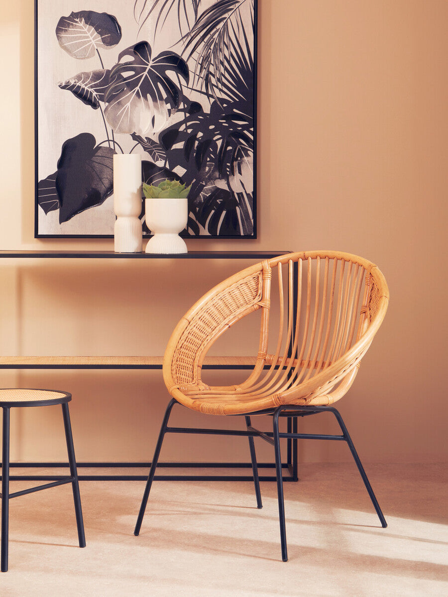 Natural rattan chair with a circular design and black iron legs, positioned next to a minimalist console table. A decorative vase and a candle sit on the table, with a large black and white artwork displayed on the wall behind. The room features warm, neutral tones