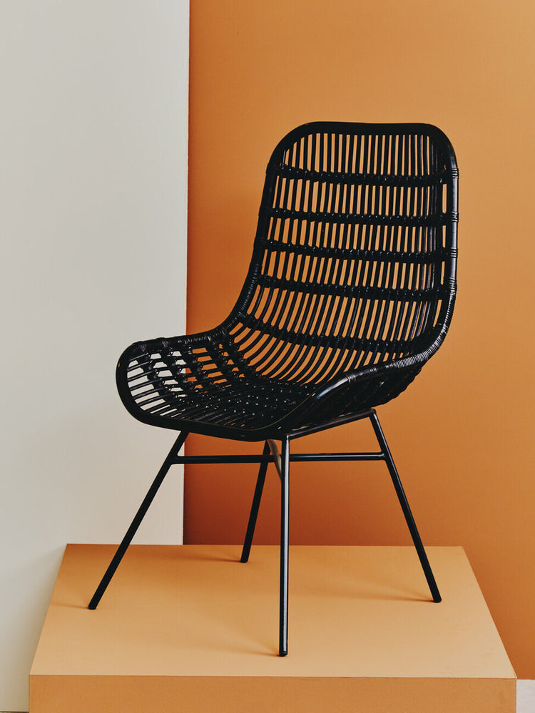 Curved black rattan chair with a natural finish, featuring a slatted design and slender metal legs, displayed against an orange backdrop. The chair has a modern aesthetic, emphasizing comfort and style