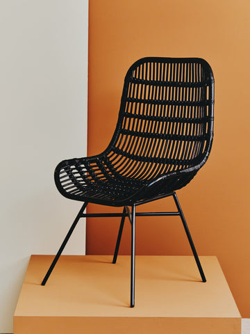 Curved black rattan chair with a natural finish, featuring a slatted design and slender metal legs, displayed against an orange backdrop. The chair has a modern aesthetic, emphasizing comfort and style