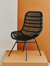Curved black rattan chair with a natural finish, featuring a slatted design and slender metal legs, displayed against an orange backdrop. The chair has a modern aesthetic, emphasizing comfort and style
