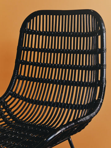 Curved black rattan chair with a slatted backrest, showcasing a natural texture against a warm orange background. The chair features a modern design with a sleek silhouette and sturdy legs