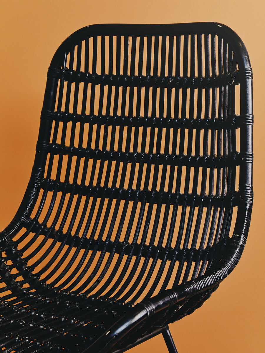 Curved black rattan chair with a slatted backrest, showcasing a natural texture against a warm orange background. The chair features a modern design with a sleek silhouette and sturdy legs