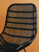 Curved black rattan chair with a slatted backrest, showcasing a natural texture against a warm orange background. The chair features a modern design with a sleek silhouette and sturdy legs