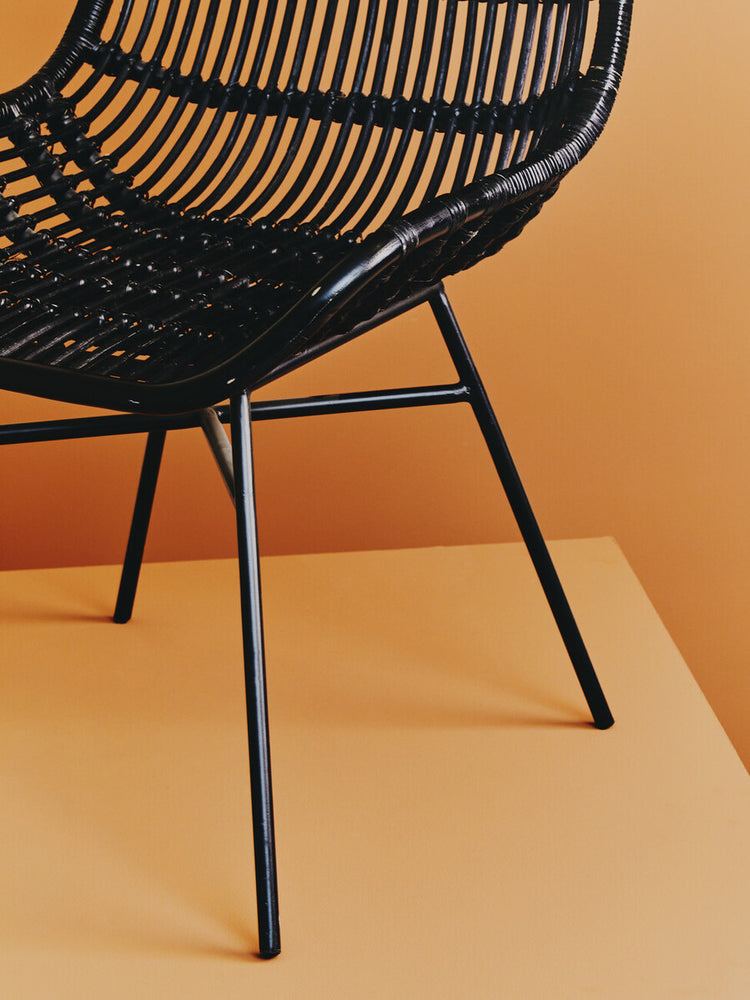 Curved black rattan chair with a natural finish, featuring a woven seat and backrest supported by sleek black metal legs, set against a warm orange background
