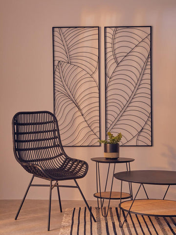 Curved black rattan chair with a sleek design, positioned next to a round wooden side table and a decorative wall art featuring leaf patterns. A small green plant is placed on the table, and a striped rug is visible on the floor. The background has a soft, neutral color