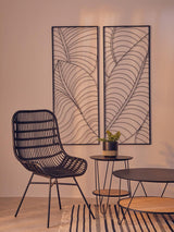 Curved black rattan chair with a sleek design, positioned next to a round wooden side table and a decorative wall art featuring leaf patterns. A small green plant is placed on the table, and a striped rug is visible on the floor. The background has a soft, neutral color