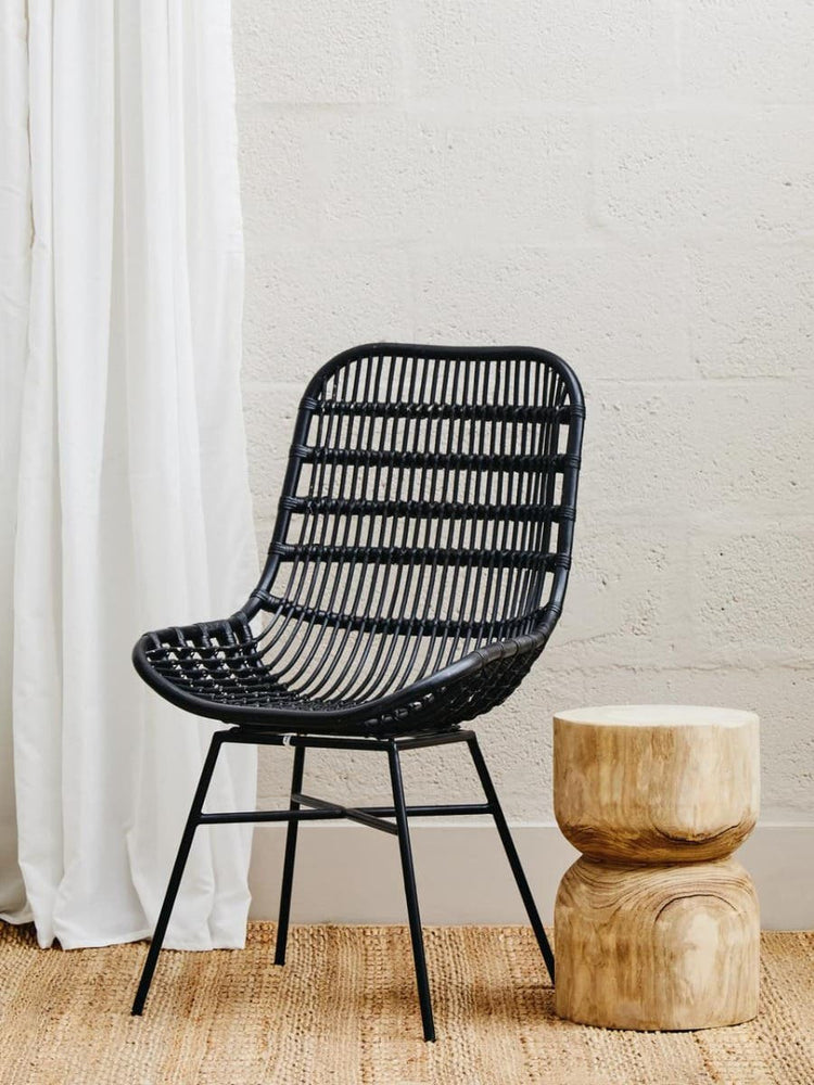 Curved black rattan chair with a slatted backrest and seat, resting on wooden legs, positioned next to two natural wood stools, against a light-colored wall and white drapery