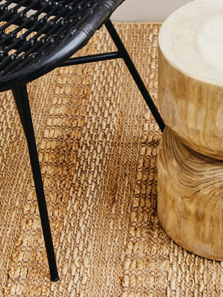 Curved black rattan chair with a sleek design, featuring thin metal legs, positioned on a textured natural fiber rug next to two round wooden stools