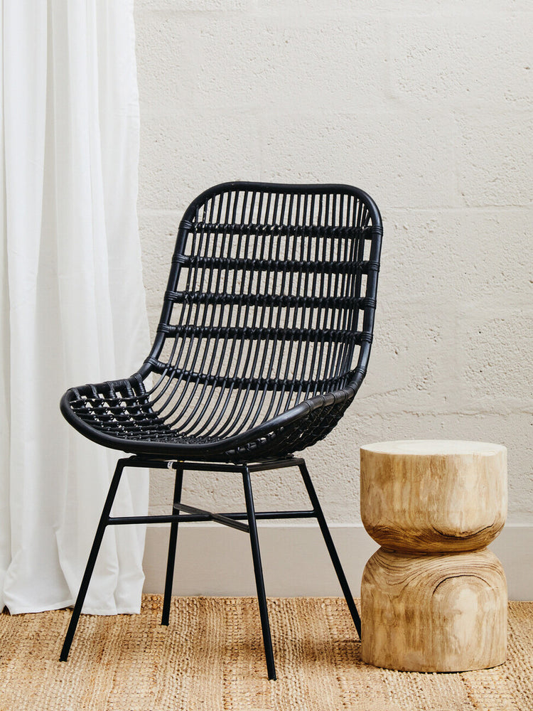 Curved black rattan chair with a slatted design, featuring a natural wood base and legs, positioned next to a round wooden side table, against a light-colored wall and draped white fabric in the background