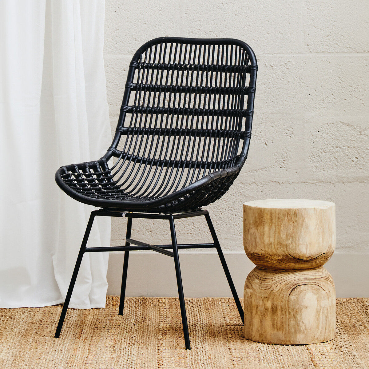 Curved black rattan chair with a slatted backrest and black metal legs, positioned next to a round wooden side table on a light wooden floor, with a neutral wall and sheer white curtain in the background