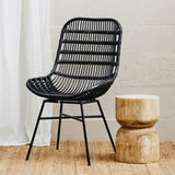 Curved black rattan chair with a slatted backrest and black metal legs, positioned next to a round wooden side table on a light wooden floor, with a neutral wall and sheer white curtain in the background