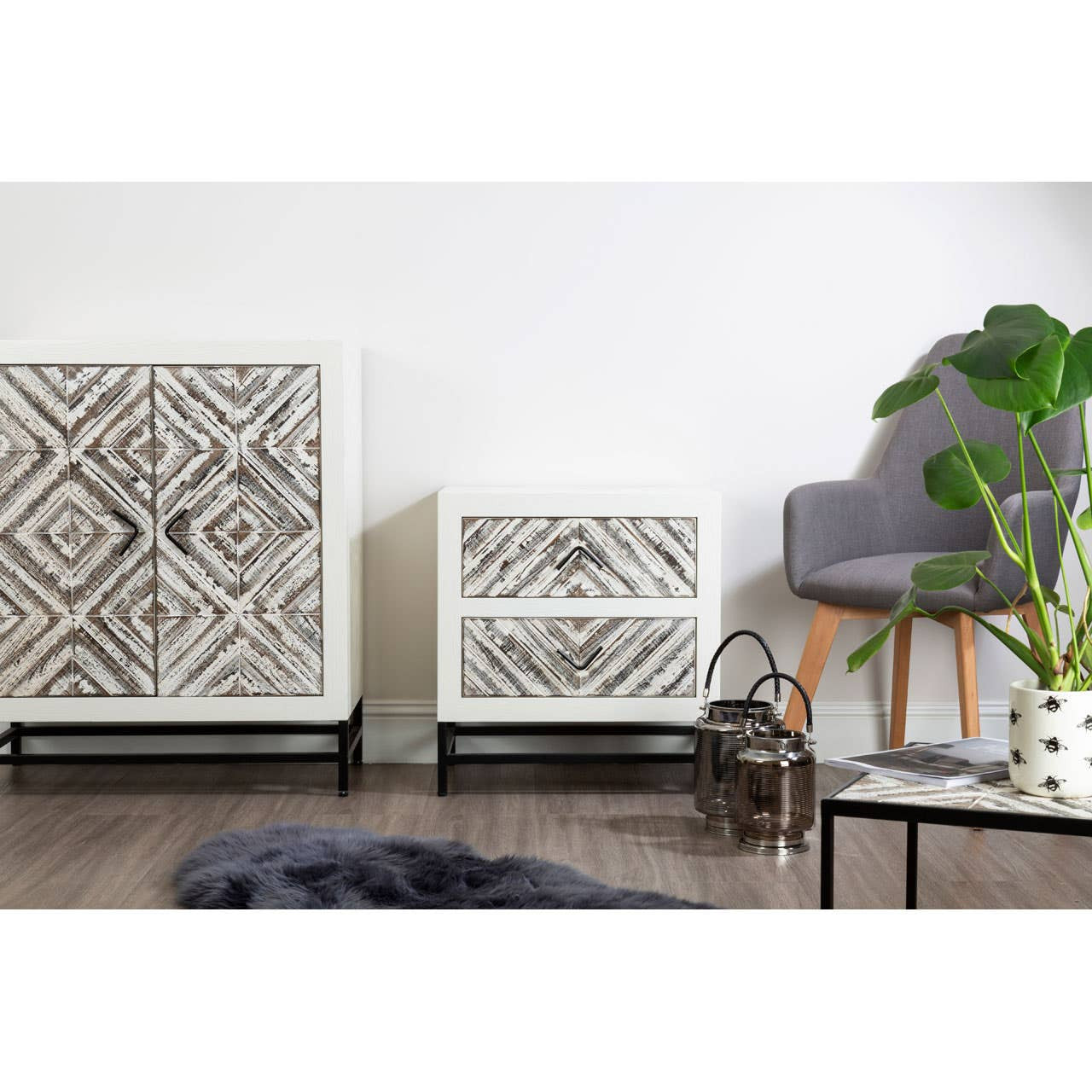 Rustic chevron-patterned two-door cabinet in a light wood finish, featuring a geometric design on the doors, set against a white wall. A smaller cabinet with a similar design is positioned beside it. A gray armchair and a potted plant are visible in the foreground, along with a black coffee table and a soft rug