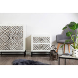 Rustic chevron-patterned two-door cabinet in a light wood finish, featuring a geometric design on the doors, set against a white wall. A smaller cabinet with a similar design is positioned beside it. A gray armchair and a potted plant are visible in the foreground, along with a black coffee table and a soft rug