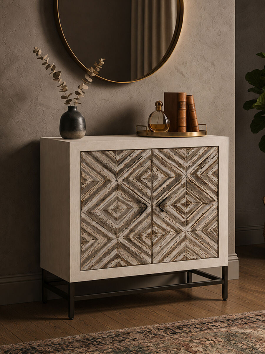A rustic two-door cabinet featuring a chevron pattern on the front, set against a neutral-toned wall. The cabinet has a light wood finish with dark metal legs, and is styled with decorative items on top, including books and a vase with branches. A round mirror hangs above the cabinet, reflecting the warm ambiance of the room. A textured rug is visible on the floor beneath the cabinet