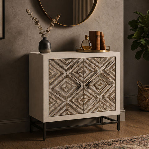 Rustic two-door cabinet featuring a chevron pattern on the front, set against a light-colored frame. The cabinet is elevated on black metal legs and includes a round mirror above it, with decorative items like a vase and books placed on top. The background has a textured wall and a hint of greenery from a nearby plant