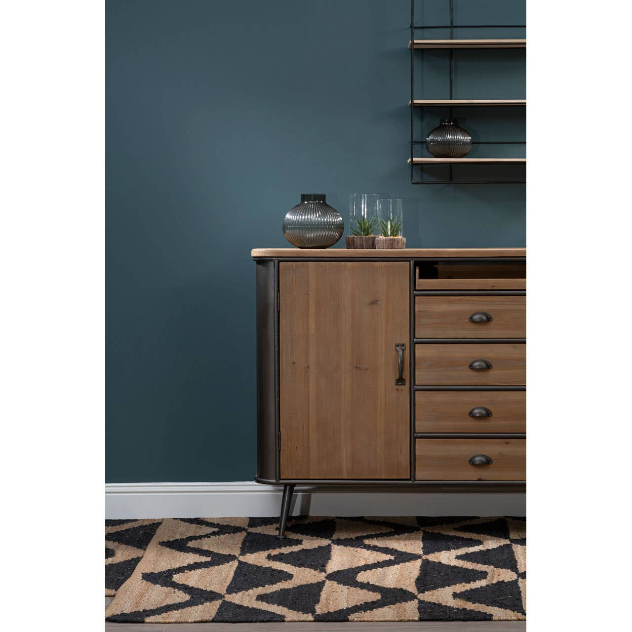 A wooden sideboard with four drawers, featuring a warm brown finish and sleek metal handles, positioned against a dark teal wall. The sideboard has an open shelf above it, displaying decorative items, and is set on a geometric patterned floor in black and beige tones