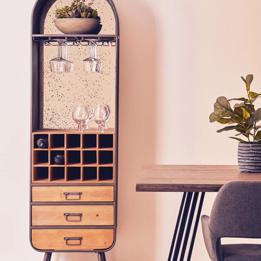 Tall wooden bar cabinet with a curved top, featuring a glass display section, wine glass holders, and multiple compartments for bottles. The cabinet has three drawers at the bottom and is positioned next to a wooden table with a potted plant on it. The background has a soft, neutral color