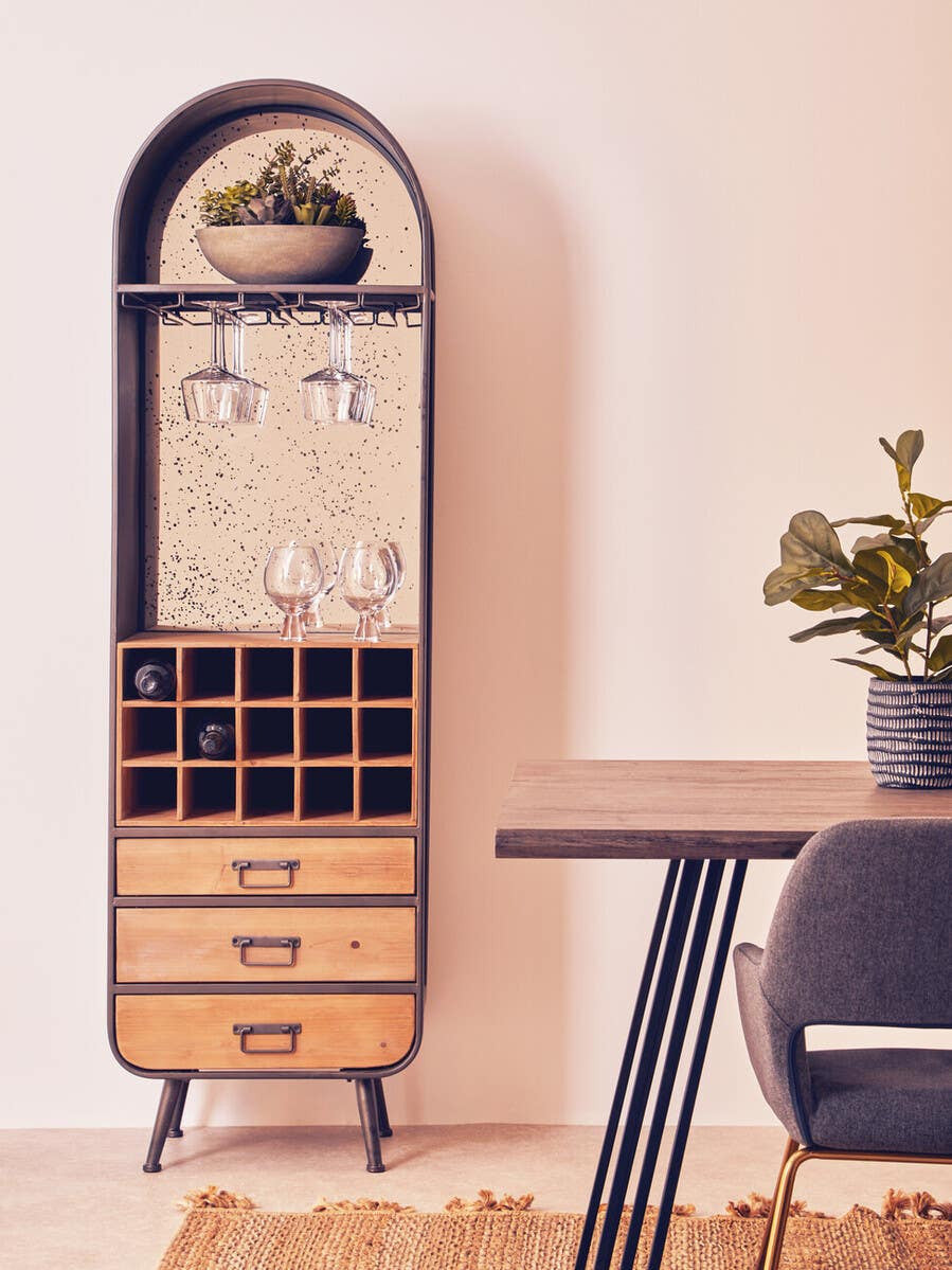 Tall wooden bar cabinet with a curved top, featuring a glass display section, wine glass holders, and multiple compartments for bottles. The cabinet has three drawers at the bottom and is positioned next to a wooden table with a potted plant on it. The background has a soft, neutral color