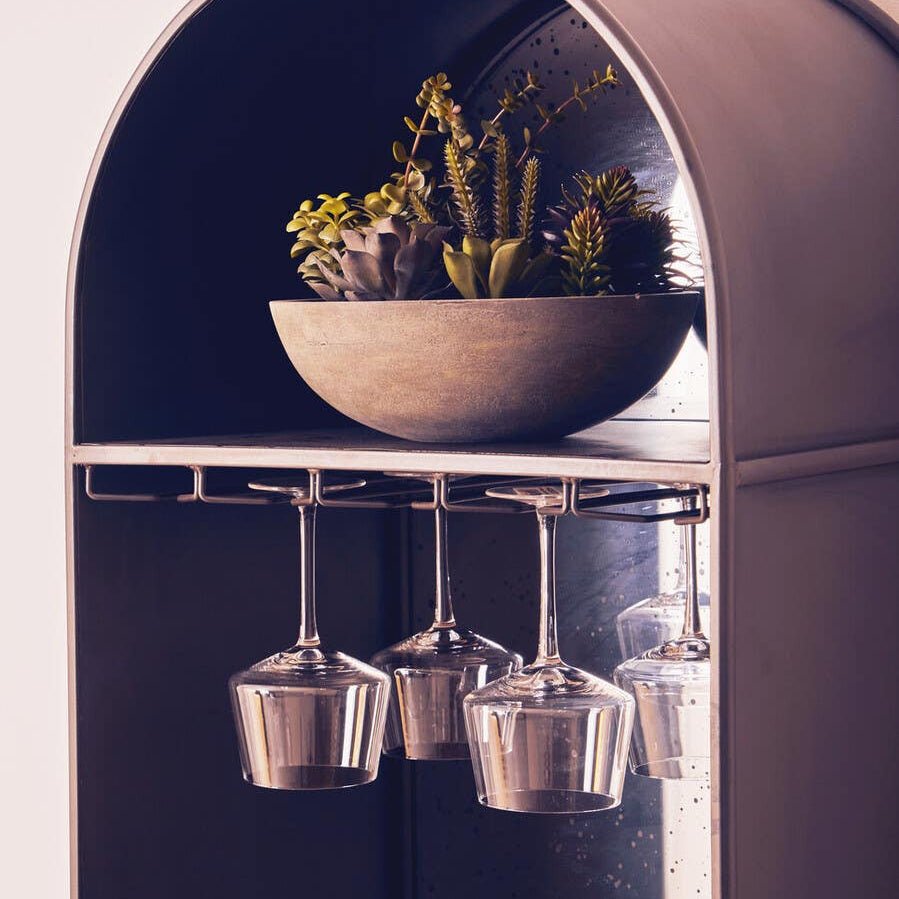 Tall wooden bar cabinet featuring an arched top, a bowl with small plants on the upper shelf, and hanging glassware below. The cabinet has a sleek design with a dark finish, enhancing its modern aesthetic