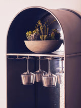 Tall wooden bar cabinet featuring an arched top, a bowl with small plants on the upper shelf, and hanging glassware below. The cabinet has a sleek design with a dark finish, enhancing its modern aesthetic