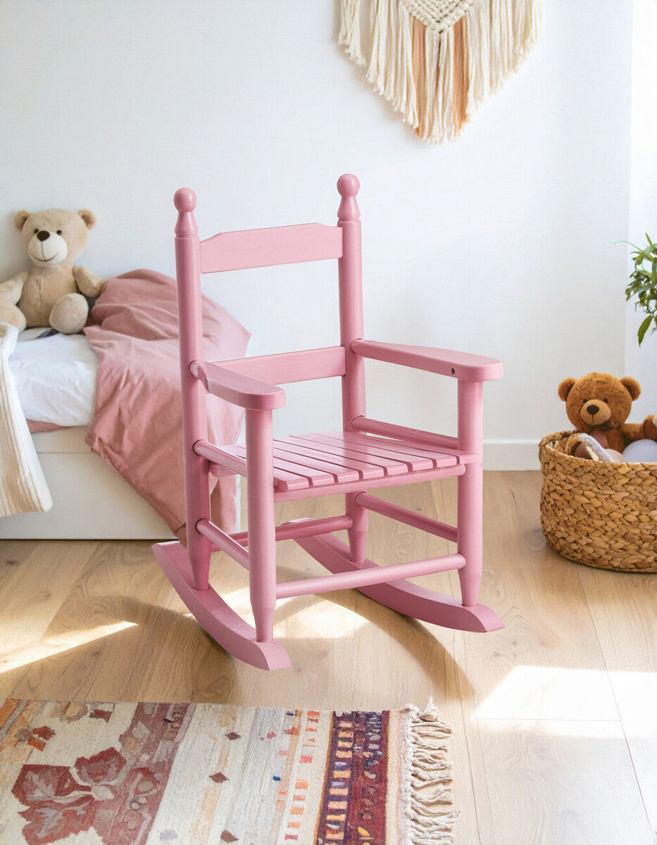 A pink rocking chair designed for kids, featuring a simple wooden structure with a high back and armrests, placed in a cozy room with a bed and decorative elements in the background. The floor is covered with a patterned rug, and a woven basket is visible nearby