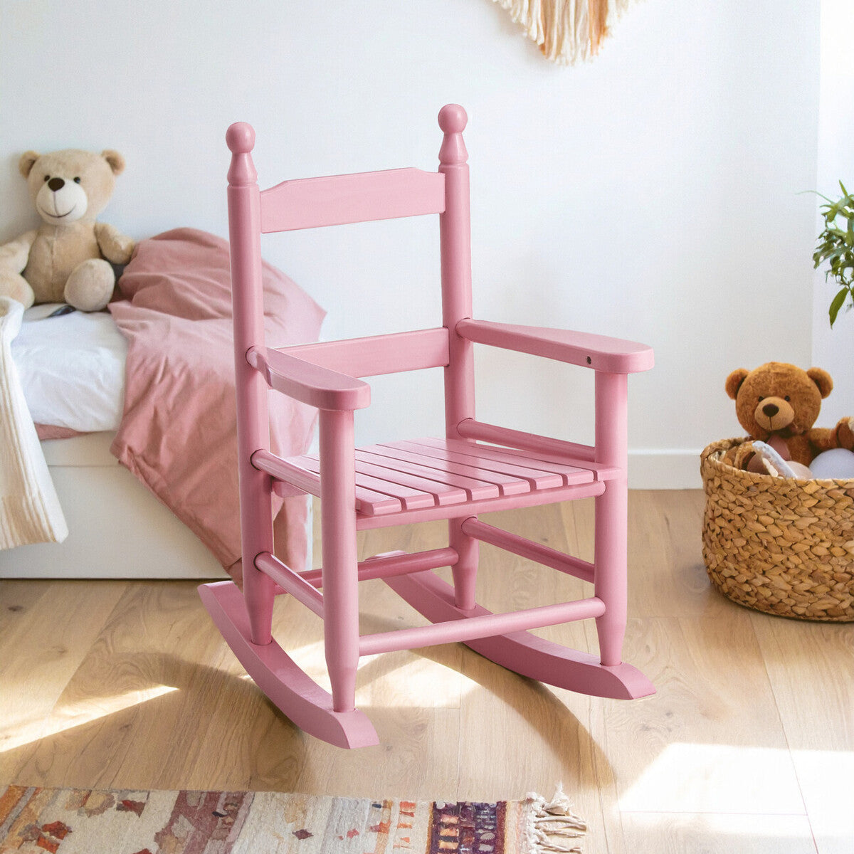 A pink wooden rocking chair designed for kids, featuring a classic style with a high back, spindles, and a sturdy seat. The chair is positioned in a bright, cozy room with a bed and soft toys visible in the background, creating a playful atmosphere