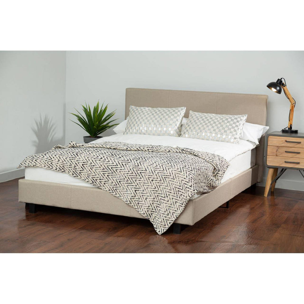Beige upholstered bed frame with headboard, zigzag throw blanket, pillows, and bedside table